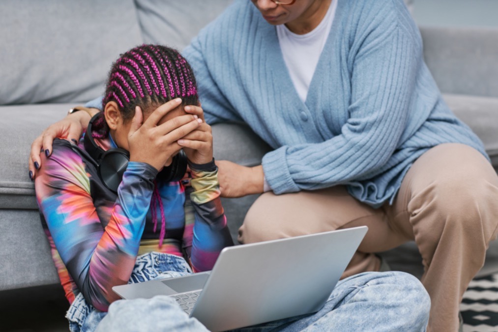 Teenager sitzt verzweifelt vor dem Laptop und wird von erwachsener Person an den Schultern berührt