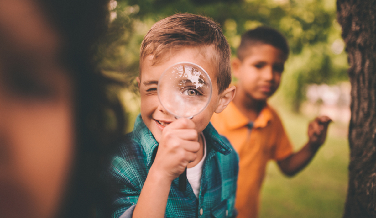 Porträt eines kleinen Jungen, der ihm eine große runde Lupe ins Gesicht hält und sein Auge humorvoll groß aussehen lässt, während er mit Freunden in einem Sommerpark spielt.