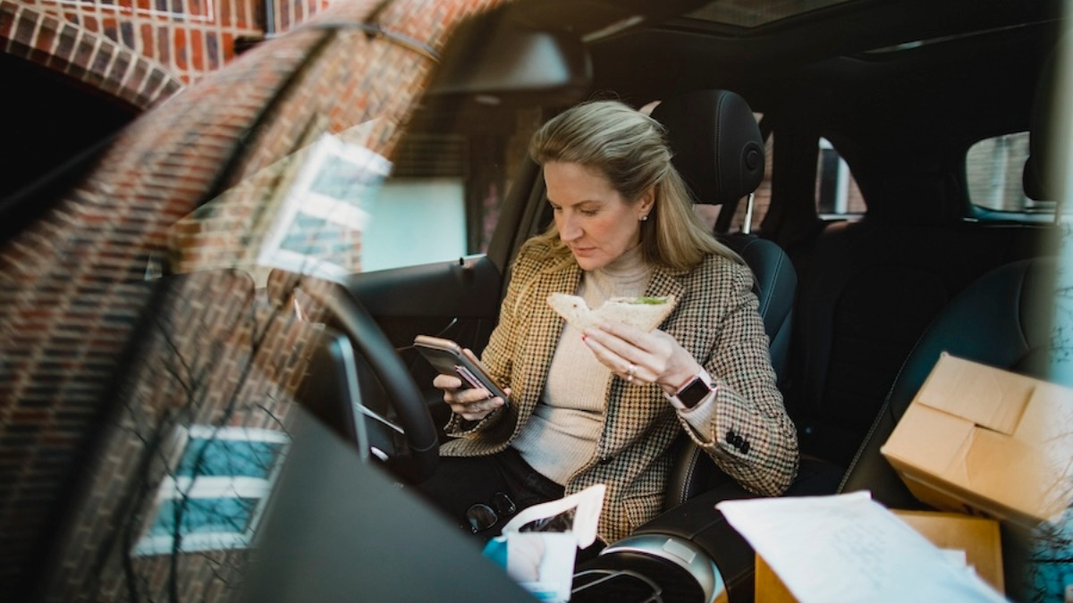 Eine Frau isst im Auto ein Sandwich und tippt dabei im Smartphone.